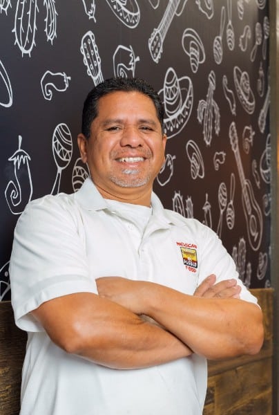 El Pueblo team member smiling, arms crossed, in front of the signature Mexican-icon mural