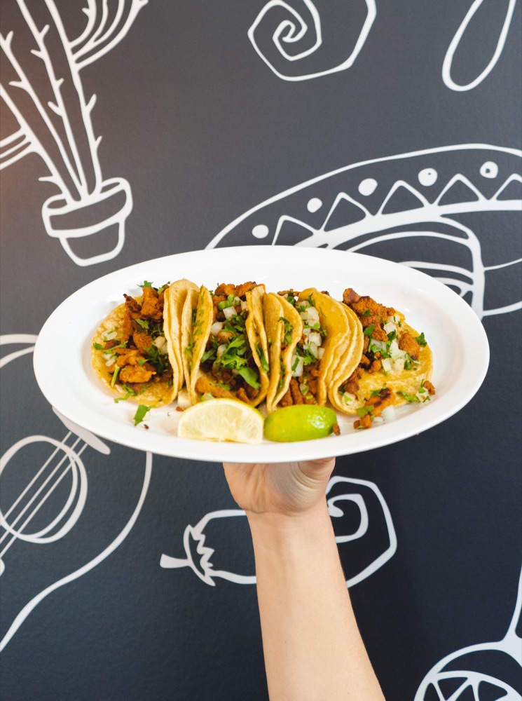 A hand holding up a plate of adobada mini tacos with onion, cilantro, and lime against the El Pueblo signature mural wall