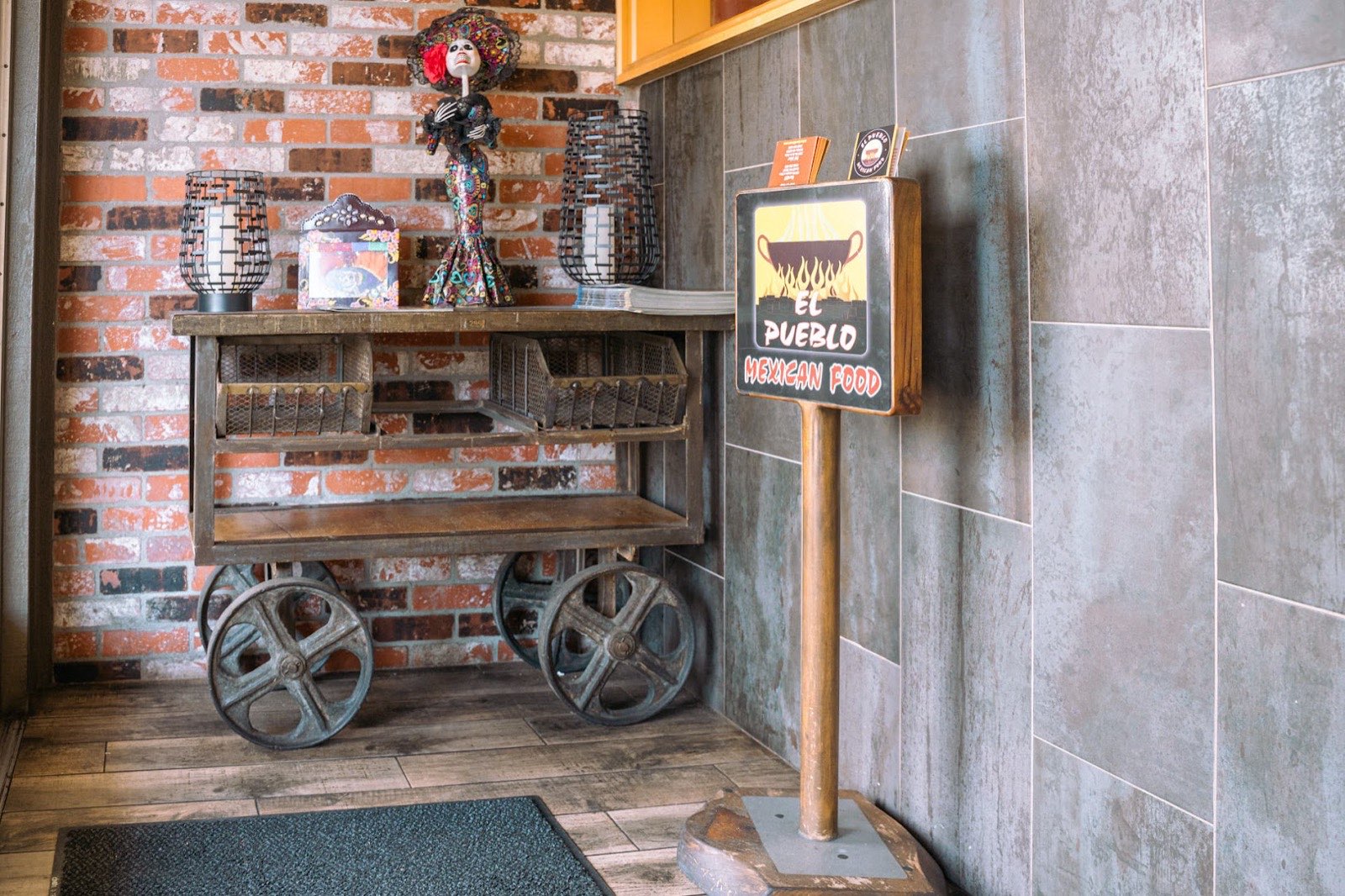 El Pueblo entryway with a Catrina figurine on a vintage cart against a brick wall, next to the menu podium